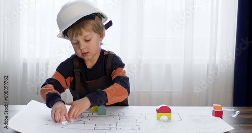 Boy with blond hair in white hardhat plays role of engineer and measures drawing with ruler. Preschooler straightens helmet and takes constructor cubes for house
