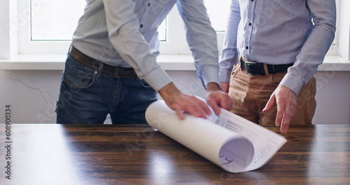 Contractors meet in spacious light office room. Worker lays out paper roll. Workmen start to have lively discussion about design of building
