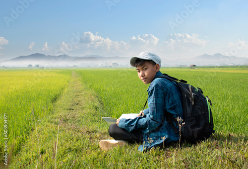 Asian boy in plaid shirt and cap sitting on paddy field in the morning and holding binoculars in hands to watch birds which flying on the sky and eating fish in rice paddy field, summer vacation.