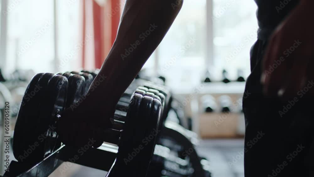 Athlete approaches the rack with sports equipment and takes a heavy dumbbell close up. Sportsman preparing to power exercise during workout in an indoor gym