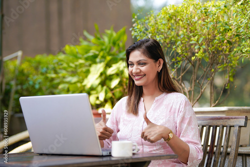 Indian woman showing thumps up while using laptop.