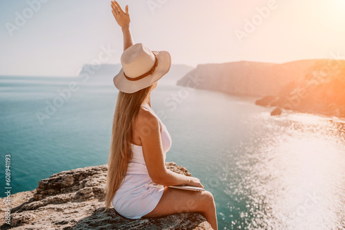 Digital nomad, woman in the hat, a business woman with a laptop sits on the rocks by the sea during sunset, makes a business transaction online from a distance. Freelance, remote work on vacation.