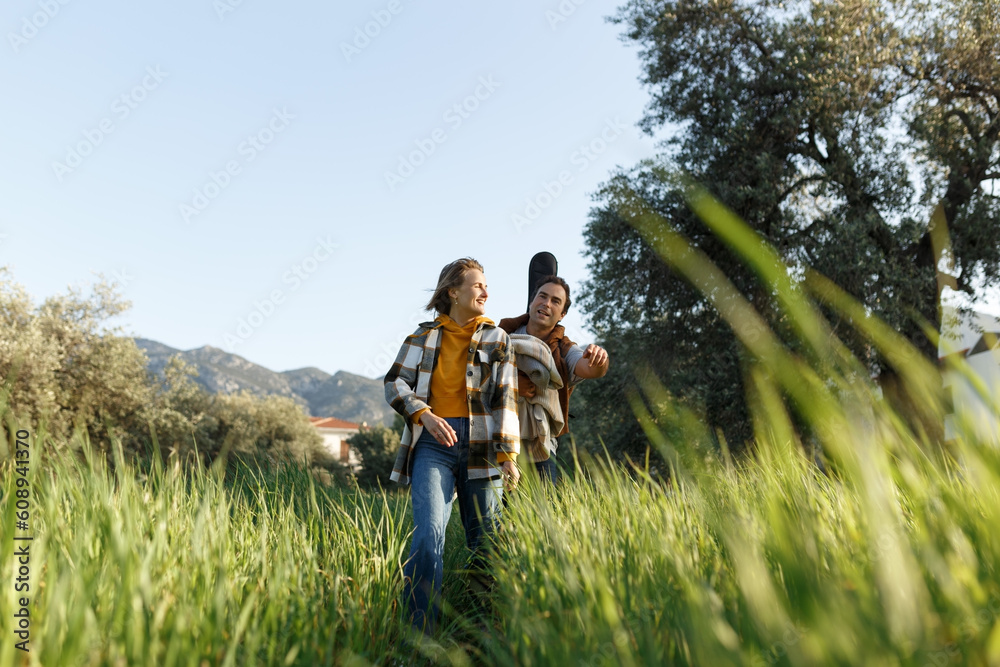 Young middle age couple walking along happily talking in park. Mature couple in summer park. Man and woman in casual clothes with guitar and picnic bag outside in spring nature. Young middle age couple walking along happily talking in park. Mature couple in summer park. Man and woman in casual clothes with guitar and picnic bag outside in spring nature.