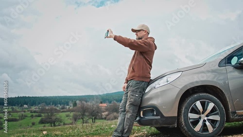 An adult man taking a picture of the view leaning against the hood of his car during a solo car trip.