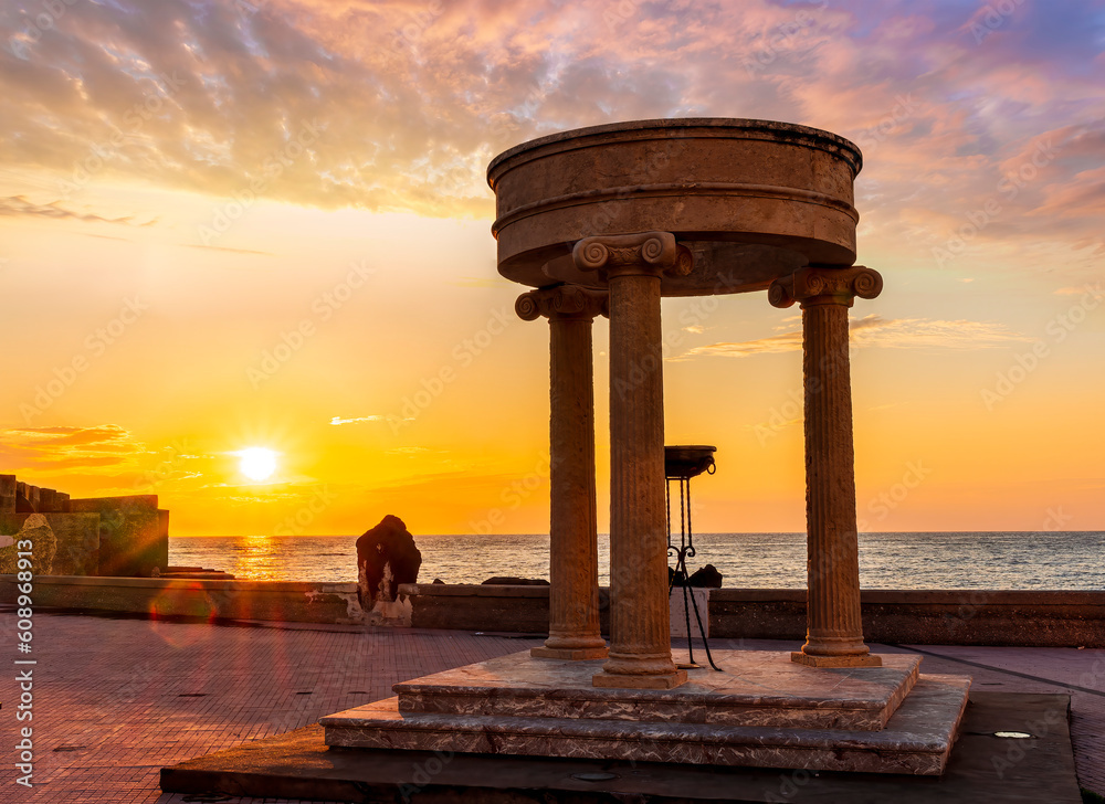 beautiful stone monument with three columns in ancient history greek style with see embarkment and amazing cloudy sunset on background of landscape