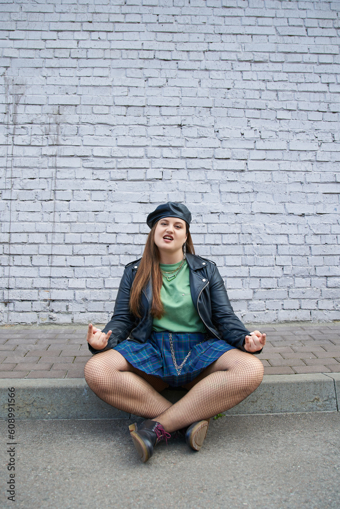 chic plus size woman posing in leather beret and jacket, plaid skirt with chains, fishnet tights and black shoes while sitting with crossed legs near brick wall on urban street, rock sign chic plus size woman posing in leather beret and jacket, plaid skirt with chains, fishnet tights and black shoes while sitting with crossed legs near brick wall on urban street, rock sign
