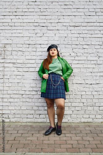 full length of brunette plus size woman posing in leather jacket with beret, plaid skirt with chains, fishnet tights and black shoes while standing near brick wall on urban street