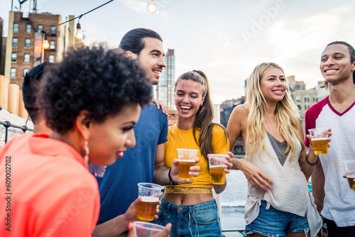 Multiethnic group of young happy friends bonding and having party at home on a rooftop terrace with New York city view