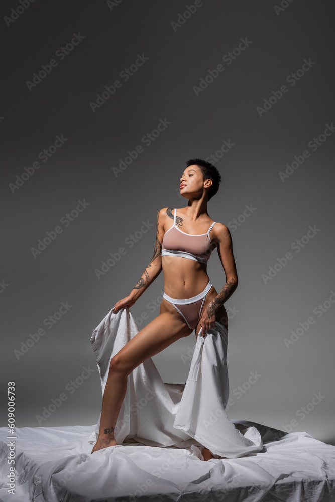 Fototapeta premium young, slender and charming woman in beige lingerie, with sexy tattooed body and short brunette hair holding bed sheet while standing on white bedding on grey background, erotic photography