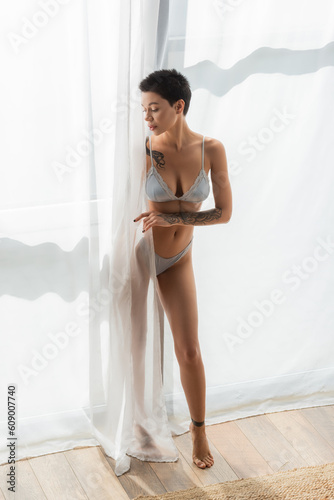 full length of young, charming and slender woman with sexy tattooed body wearing grey silk lingerie while standing in natural light near white curtain in modern bedroom at home © LIGHTFIELD STUDIOS