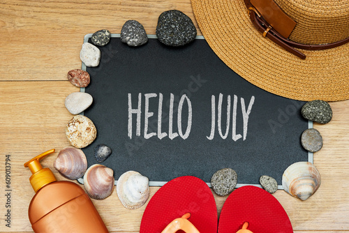 summer concept, sun cream and slipper with hello July text on chalkboard and stones. seashell hat on wood table
