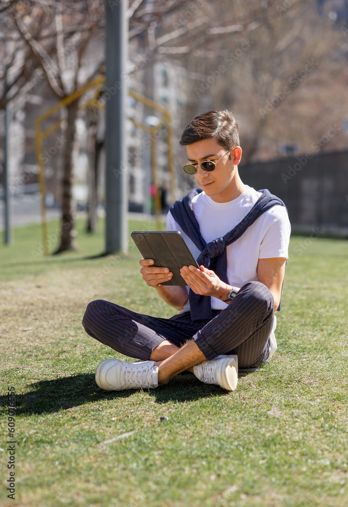 Fototapeta premium a young man is sitting with a tablet in his hand in a park, concept of remote work, online communication, using social networks, studying or working outdoors