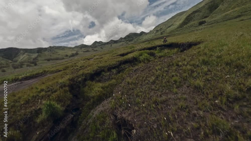 Aerial view biker riding on moto enduro path between autumn grass volcano mountain landscape extreme. FPV drone follow shot motorbike motocross speed racing power race trip at rock scenery cloudy sky