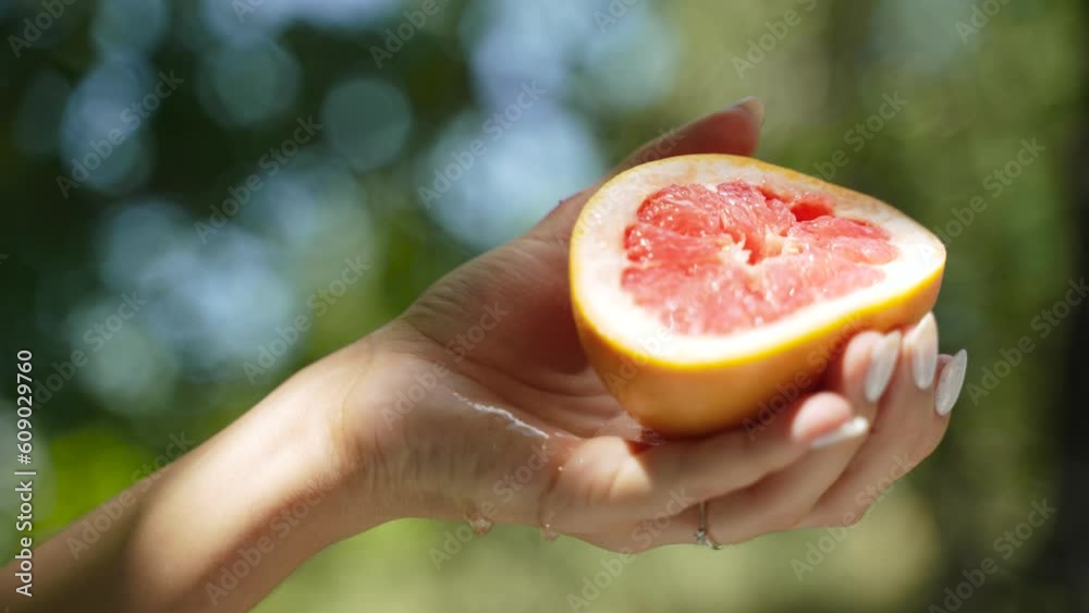 Piece of juicy grapefruit is squeezed out by graceful female hand with white manicure in garden in sunlight. Citrus juice flows down palm of hand, on hand is shadow from branches of trees.