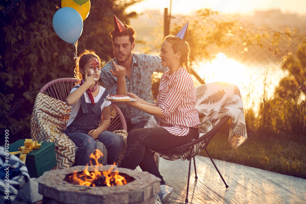 Happy family celebrating birthday of daughter outdoors in nature, sitting by fireplace, trying cake, birthday girl with cake on her face. Happy family celebrating birthday of daughter outdoors in nature, sitting by fireplace, trying cake, birthday girl with cake on her face.