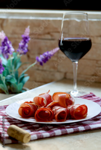Plate with sliced prosciutto and a glass of red wine on kitchen table, lifestyle photography. Prsut and red wine, italian or spanish cured meats. Jamon appetizer with a glass of wine, brunch concept
