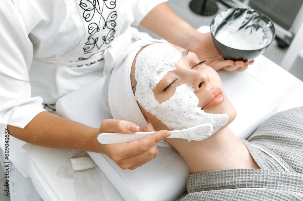 Obraz premium Close-up face of a beautiful woman lying with her eyes closed in the salon, beautician applies a moisturizing anti-aging mask on her face. Skin care, beauty procedures, spa day