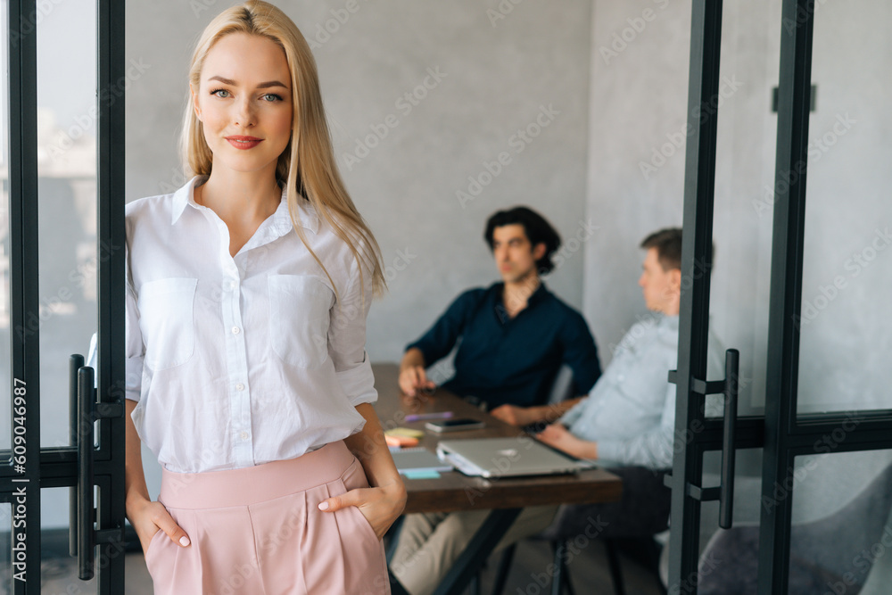 Obraz premium Portrait of pretty blonde businesswoman in casual clothes standing in modern office room, confident looking at camera. Startup business team discussing project sitting at desk on background