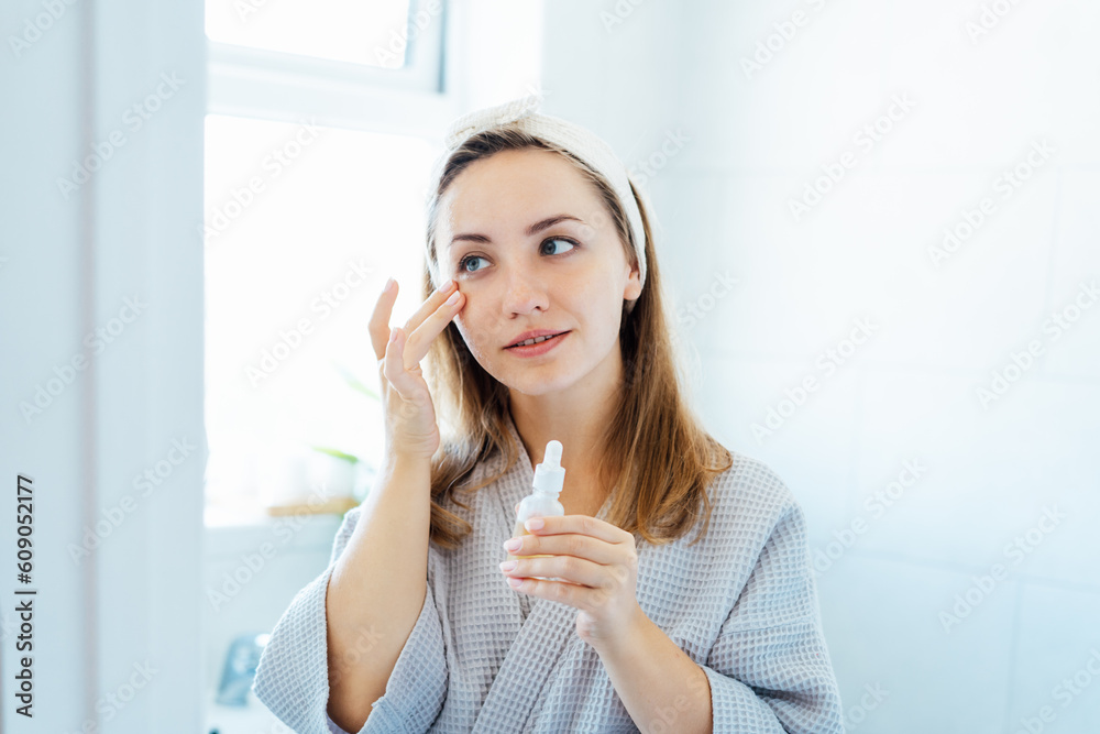 Young woman in bathrobe looking in the mirror, holding bottle with hyaluronic acid serum and applying it on face. Skin hydrating. Cosmetic spa procedures. Beauty self-care at home. Selective focus Young woman in bathrobe looking in the mirror, holding bottle with hyaluronic acid serum and applying it on face. Skin hydrating. Cosmetic spa procedures. Beauty self-care at home. Selective focus