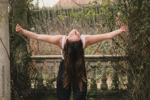 Female yogi in modified standing back bend with eyes closed in a leafy terrace of a landscaped garden with stone columns and balustrade. Brunette long hair. Outdoor yoga practice. Gyan Mudra hands.