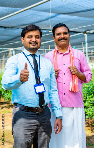 Vertical shot of happy smiling indian banker or officer showing thumbs up sign with farmer by looking camera in at greenhouse - concept of approval, encouragement and authorization