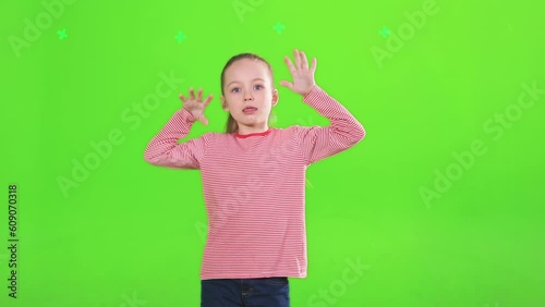 Funny adorable girl making funny faces, while looking at camera in studio. Pretty female kid teasing, pulling tongue out, raising hands, isolated on green, in motion. Concept of expression, teasing.