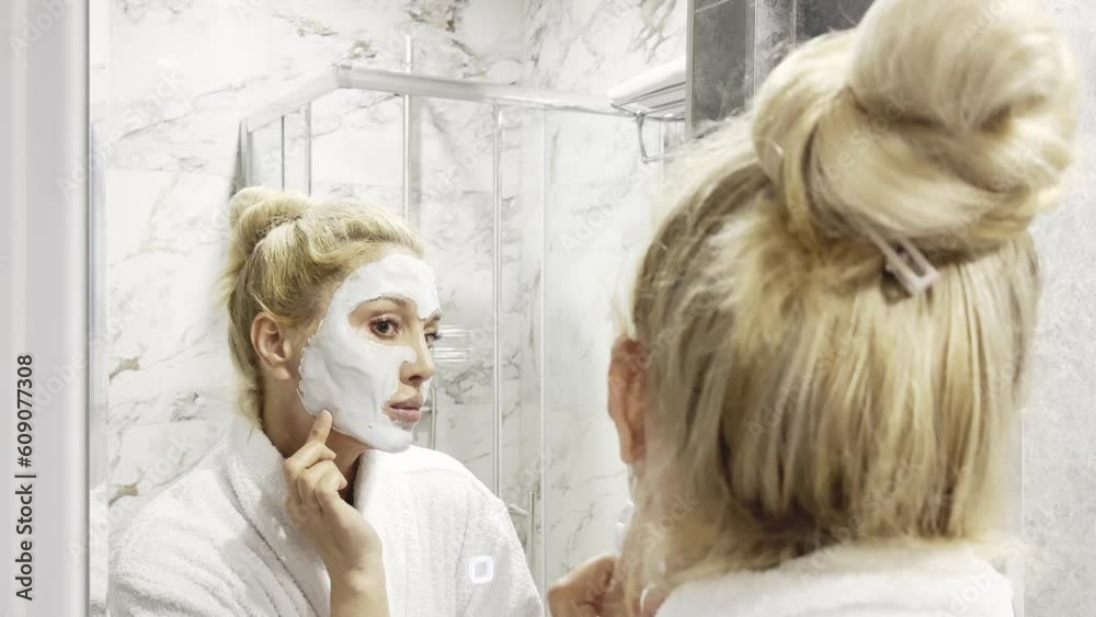 Woman in bathroom using skin mask and looking in mirror Woman in bathroom using skin mask and looking in mirror