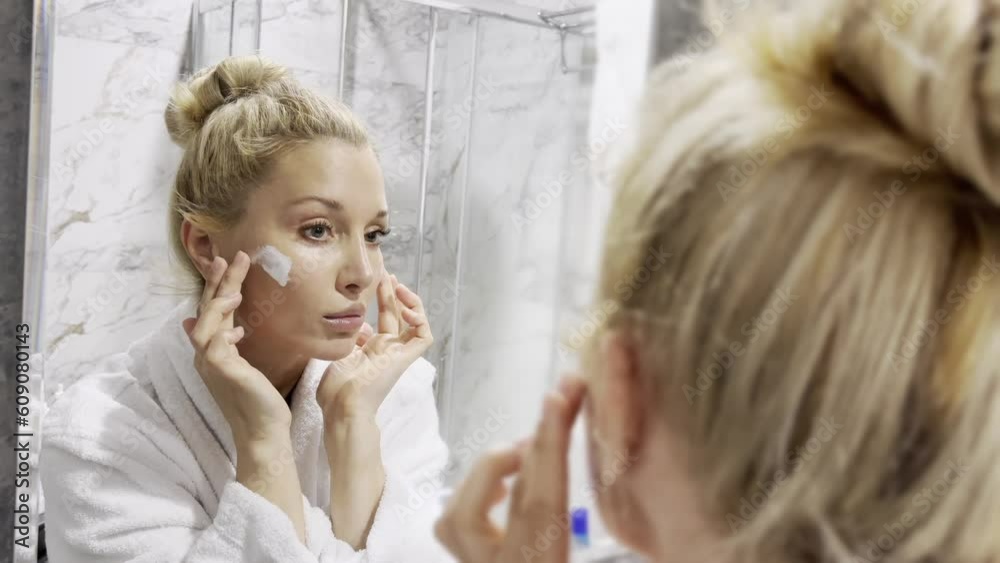 mid age adult blonde woman wears bathrobe and towel in bathroom applying rejuvenating tightening antiage face skin care cream treatment. Daily beauty routine. mid age adult blonde woman wears bathrobe and towel in bathroom applying rejuvenating tightening antiage face skin care cream treatment. Daily beauty routine.