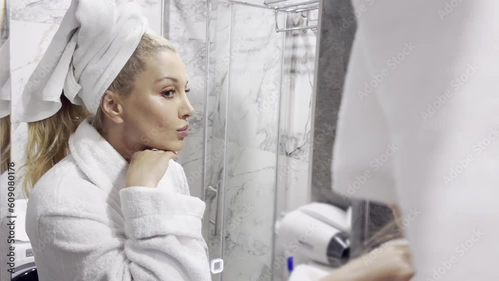 mid age adult blonde woman wears bathrobe and towel in bathroom applying rejuvenating tightening antiage face skin care cream treatment. Daily beauty routine. mid age adult blonde woman wears bathrobe and towel in bathroom applying rejuvenating tightening antiage face skin care cream treatment. Daily beauty routine.