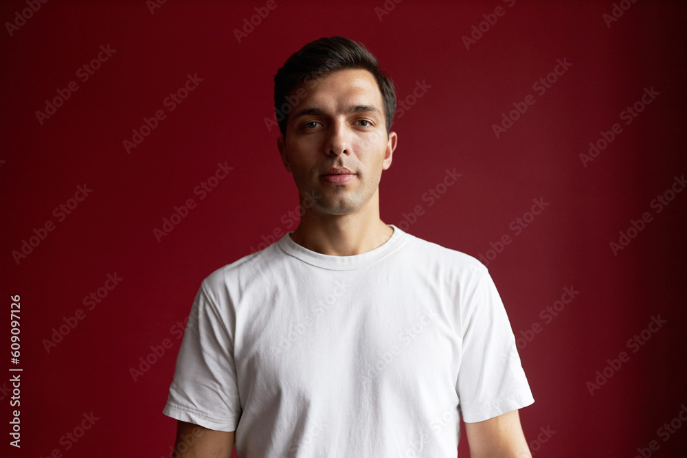 Waist up front view portrait of youthful guy in white mockup t-shirt with copy space for your commercial or promo content, standing against red studio background, looking straight at camera Waist up front view portrait of youthful guy in white mockup t-shirt with copy space for your commercial or promo content, standing against red studio background, looking straight at camera