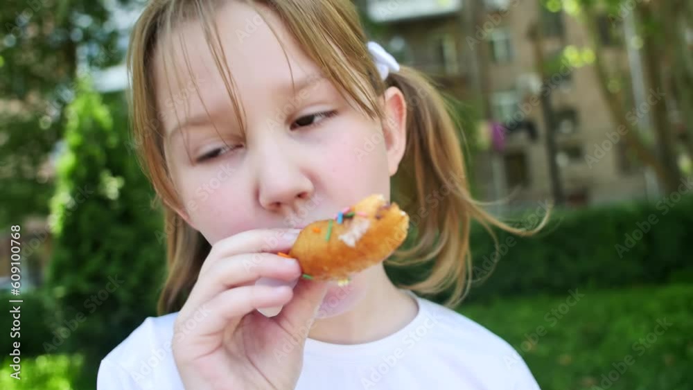 A cute baby girl in a white T-shirt with pigtails and bows on her hair bites off a donut and eats outdoors smiling cheerfully playfully. A snack for a teenager on a walk in the summer. A cute baby girl in a white T-shirt with pigtails and bows on her hair bites off a donut and eats outdoors smiling cheerfully playfully. A snack for a teenager on a walk in the summer.