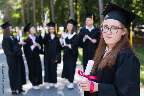 Fototapeta Portrait of a young caucasian woman in glasses and a graduate gown against the background of classmates