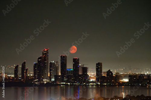 An early morning view of the moon setting behind buildings. The moon is a deep red because of smoke from wildfires in western Canada