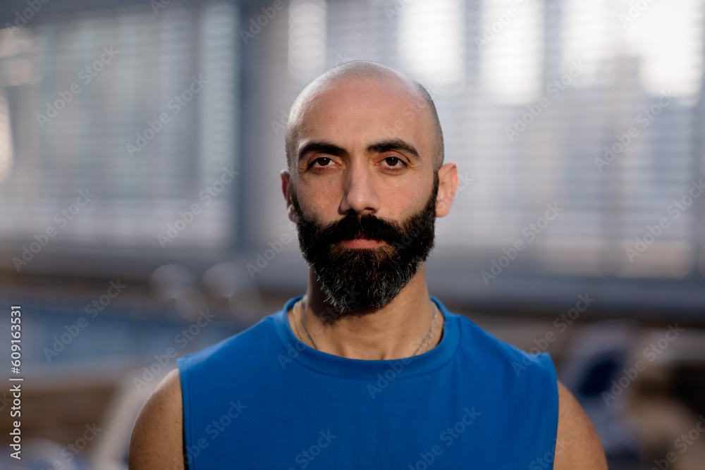 Standing by a poolside, a man in athletic wear poses for a portrait with dramatic golden hour light. He has a shaved head, a gray beard flecked with gray, and a sleeveless blue shirt. Standing by a poolside, a man in athletic wear poses for a portrait with dramatic golden hour light. He has a shaved head, a gray beard flecked with gray, and a sleeveless blue shirt.