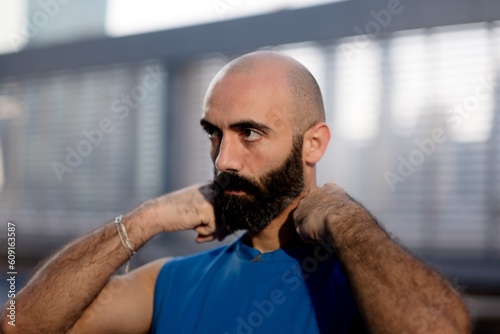 Adjusting his collar, an athletic man with a shaved head, black beard, and hairy arms stands outside by a pool. photo