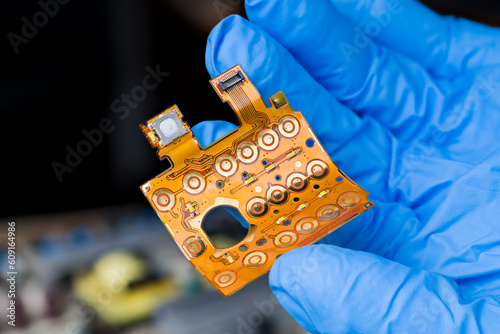 Closeup a flexible printed circuit of keypad membrane on engineer hand in blue glove. Round button switches, LED diodes or flex flat cable with connector on bent plastic board on dark blur background.