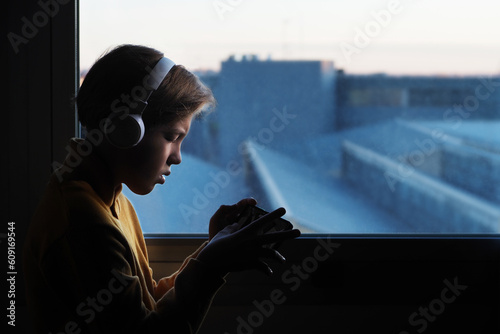 Horizontal photo of a 12-year-old teenager sits by the window overlooking the city, wearing white headphones and looking at their phone