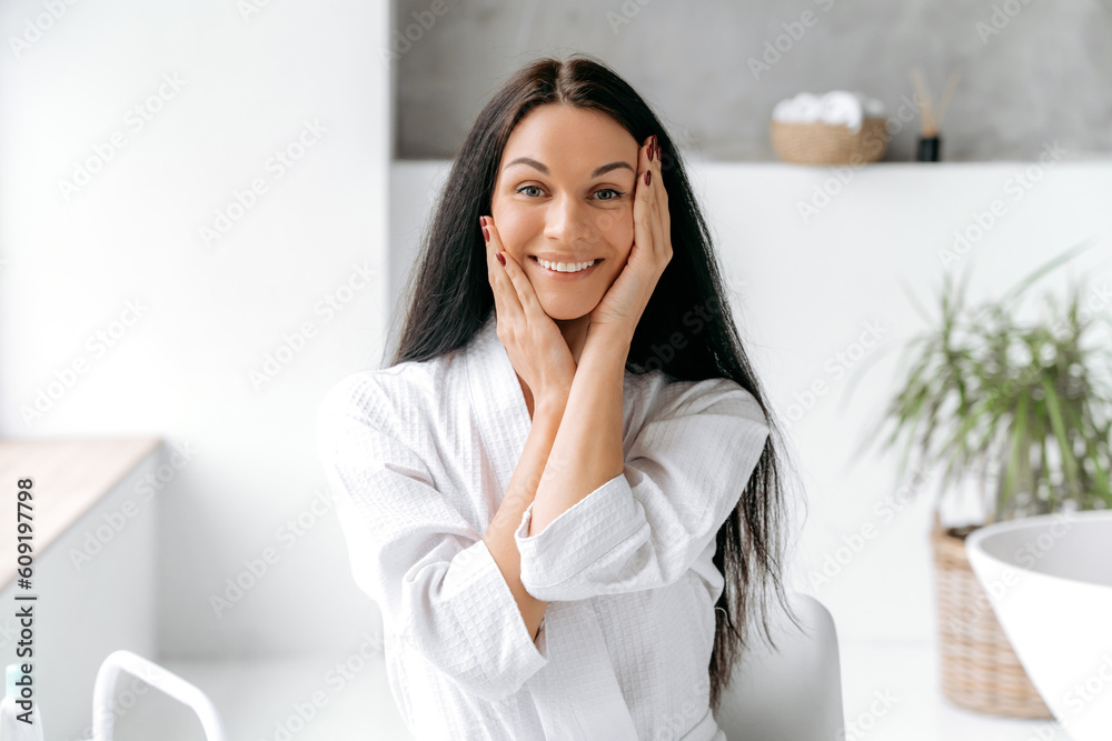 Photo of a beautiful well-groomed caucasian young woman with long black hair, in a bathrobe, stand in the bathroom, posing holding her hands near her face, looking at the camera, smiling happily Photo of a beautiful well-groomed caucasian young woman with long black hair, in a bathrobe, stand in the bathroom, posing holding her hands near her face, looking at the camera, smiling happily