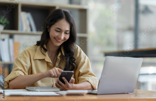 Asian businesswoman using mobile phone to search information and chat via application while sitting at work using laptop with paperwork making plan, investing, analyzing financial report.