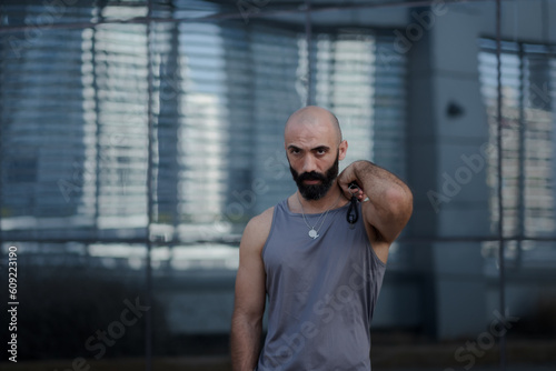 With one hand on his shoulder, a man poses for a portrait in a sleeveless shirt. He is wearing athletic wear, and has a toned physique. A touch of gray in his beard expresses he is a middle-aged man. photo