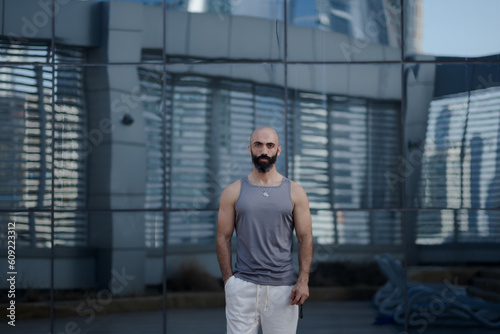 An athletic man in a gray sleeveless shirt and white sweat pants poses in front of a building's glassy, reflective facade. He has toned arms and a graying beard, showing fitness in middle age.