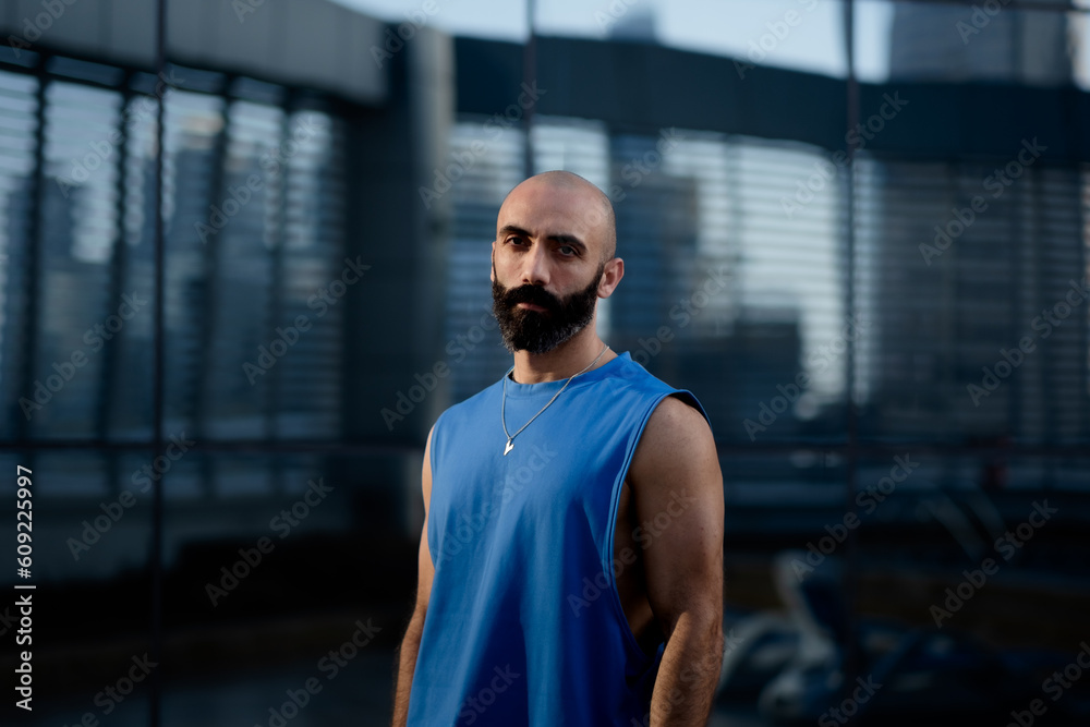 A man in a blue sleeveless shirt poses in warm, golden sunlight that accentuates his toned physique and thick beard. In the background, a reflective glass wall shows an outdoor fitness area.