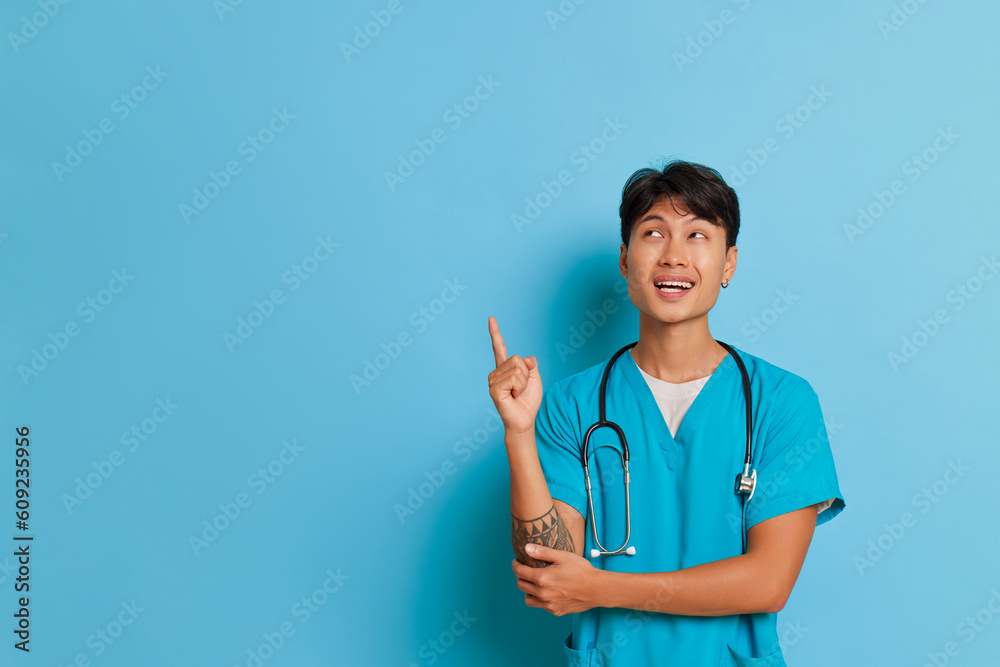 Young male doctor stands on blue background, smiling and pointing with index finger up at empty space, business concept, copy space Young male doctor stands on blue background, smiling and pointing with index finger up at empty space, business concept, copy space