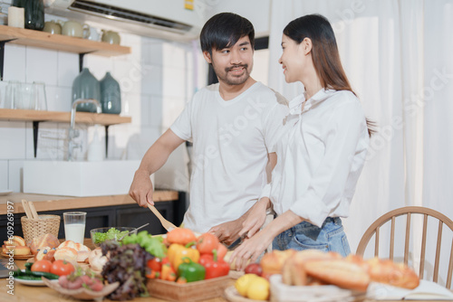 Smiling man hugging woman, two people standing and joyfully looking at each other. Young couple happily spending time in cozy modern kitchen at home.