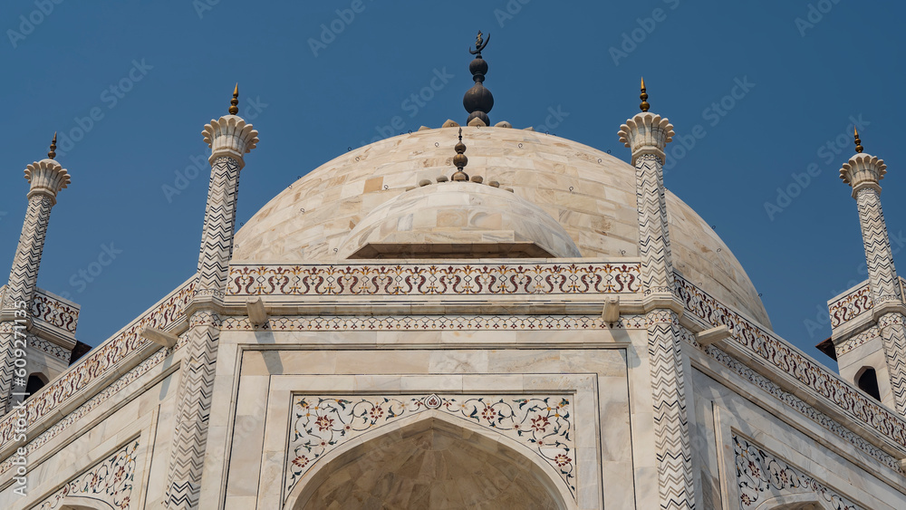 The upper part of the Taj Mahal mausoleum against the blue sky. White marble dome with spires. There are ornaments on the walls, inlays of precious stones with floral motifs. Close-up. India. Agra
