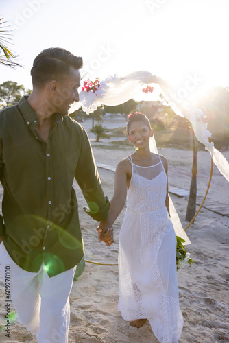 Happy caucasian newlywed couple holding hands and walking at beach at wedding ceremony at sunset