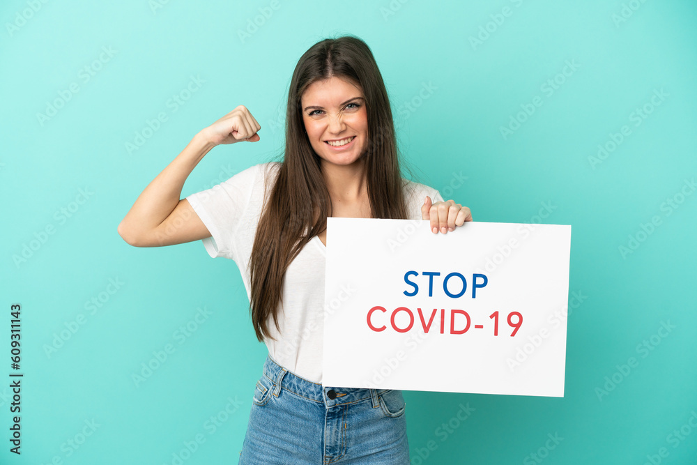 Young caucasian woman isolated on blue background holding a placard with text Stop Covid 19 and doing strong gesture