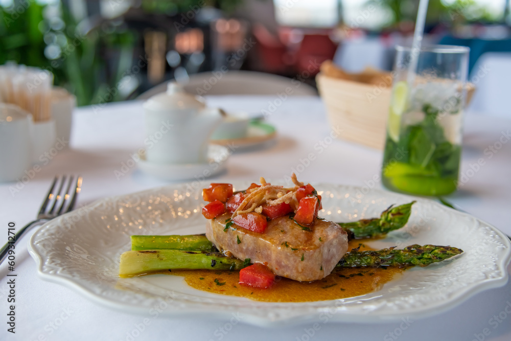 Close-up view of fried tuna steak with grilled asparagus, tomatoes and salt oyster sauce on white plate in seafood restaurant. Soft focus. Gourmet theme. Close-up view of fried tuna steak with grilled asparagus, tomatoes and salt oyster sauce on white plate in seafood restaurant. Soft focus. Gourmet theme.