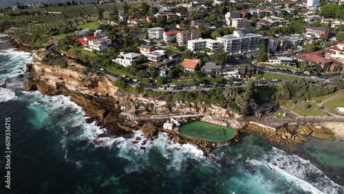 Zoom out/reveal shot of oceanfront property looking over Bronte Baths ocean pool with waves crashing against rocks from above. Drone 4K.