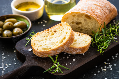Ciabatta bread on wooden board with olive oil, olives and herbs on black. Mediterranean food.